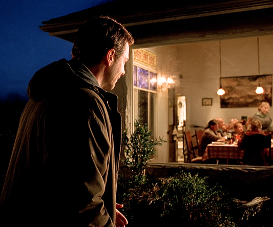 A man standing outside at night looking through the window of a lit room where a family sits at a table